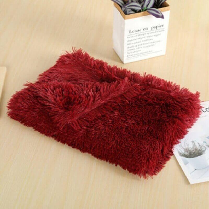 ワインレッドのふんわり毛布がきれいにたたまれて、木のテーブルの上に。そばに小さな観葉植物と書類があるよ